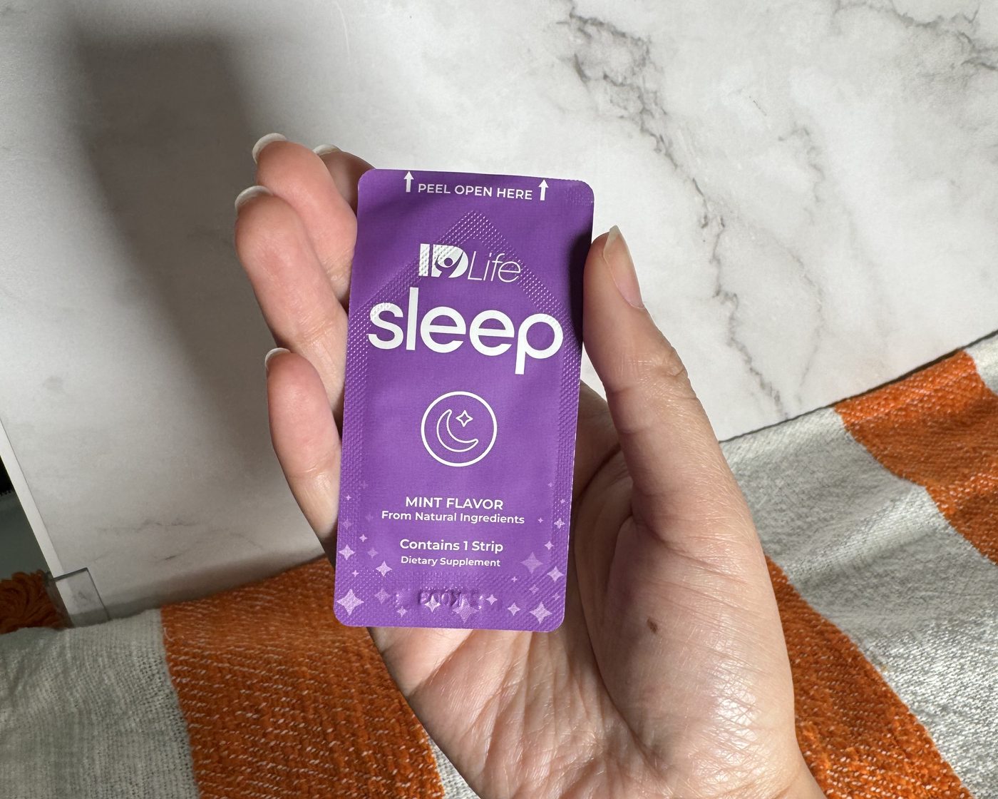 A hand holding one purple IDLife Sleep strip pack over an orange and white striped blanket