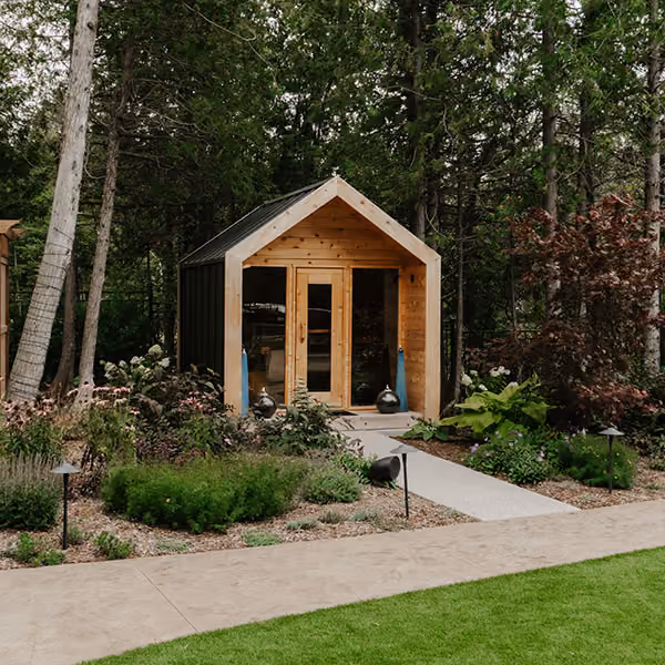 Small modern wooden cabin in a garden surrounded by lush trees and plants with a paved pathway.