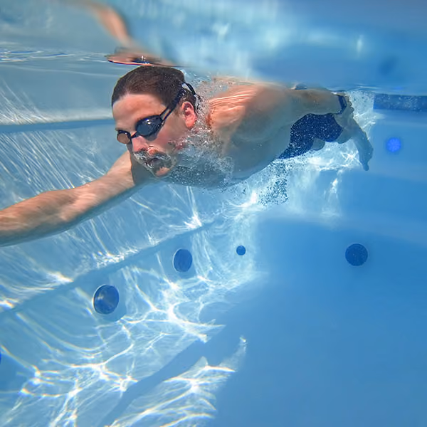 Man wearing goggles swimming underwater in a clear pool with blue tiles and lights.