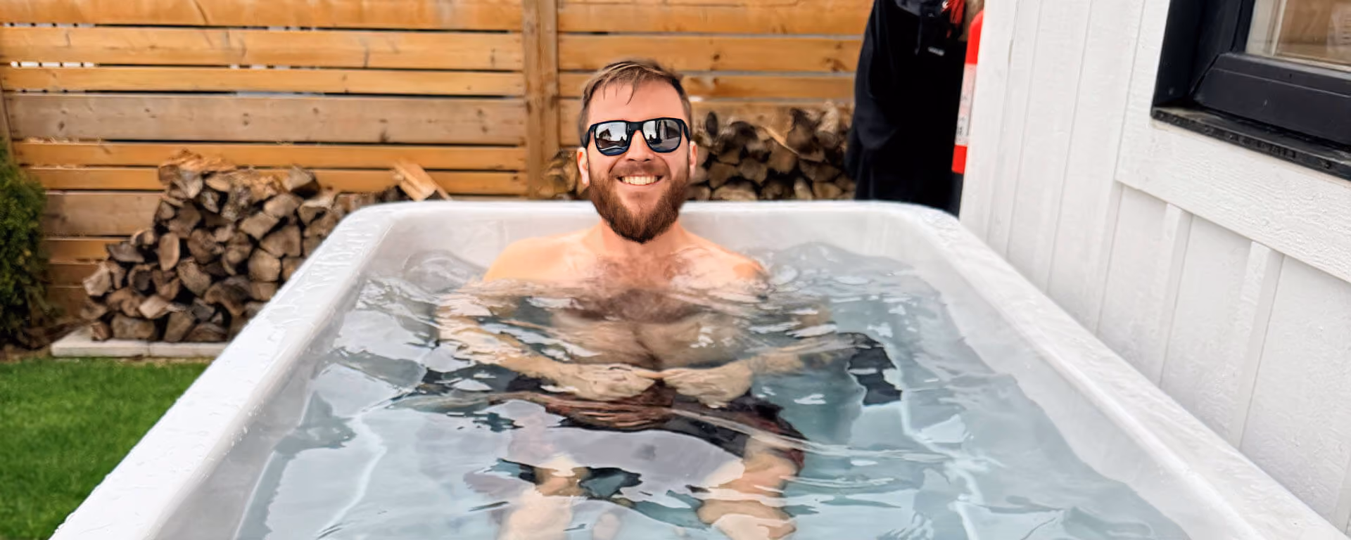 Bearded man wearing sunglasses smiling while relaxing in an outdoor cold plunge tub.