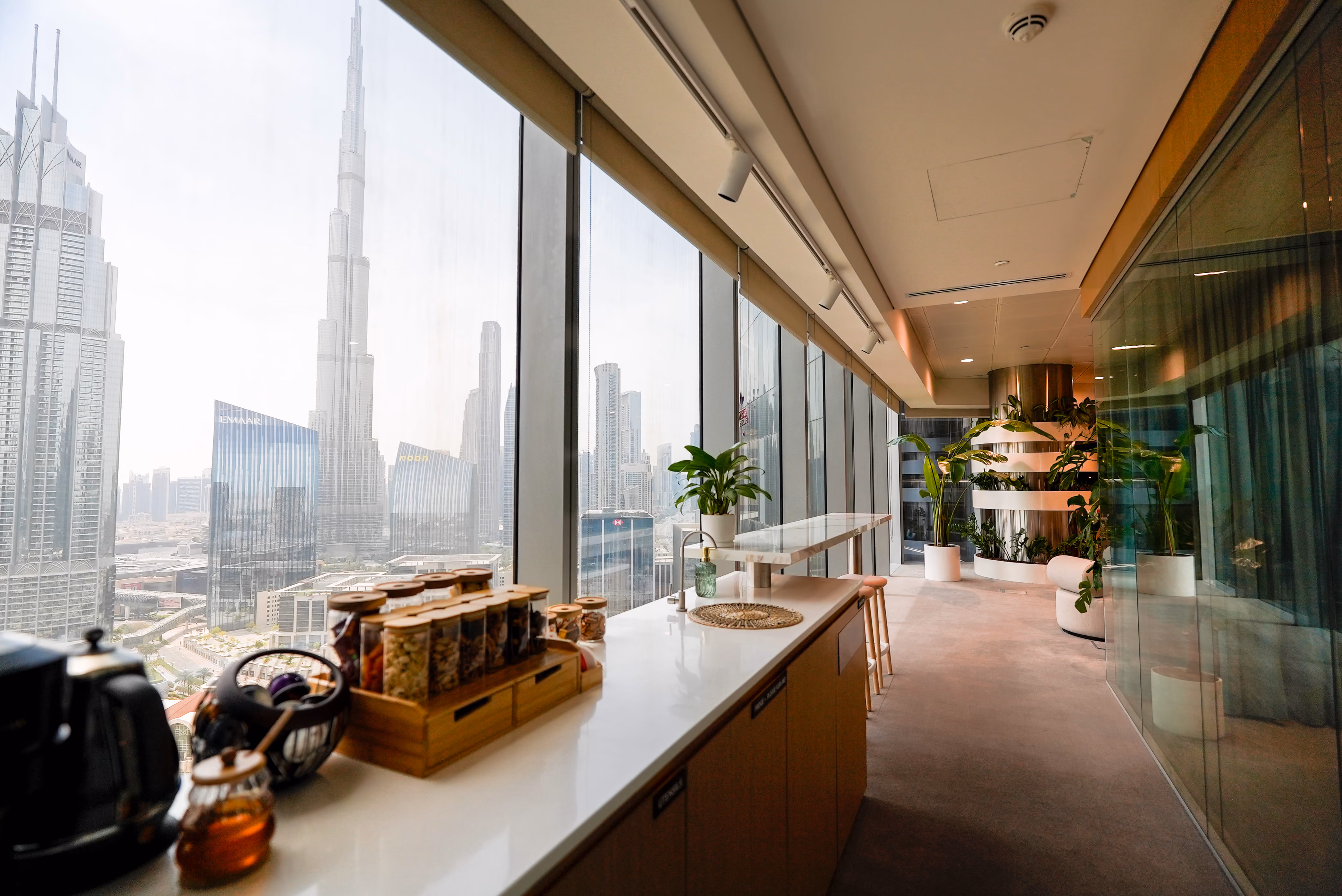 Modern office kitchen with floor-to-ceiling windows showing Dubai skyline and Burj Khalifa.