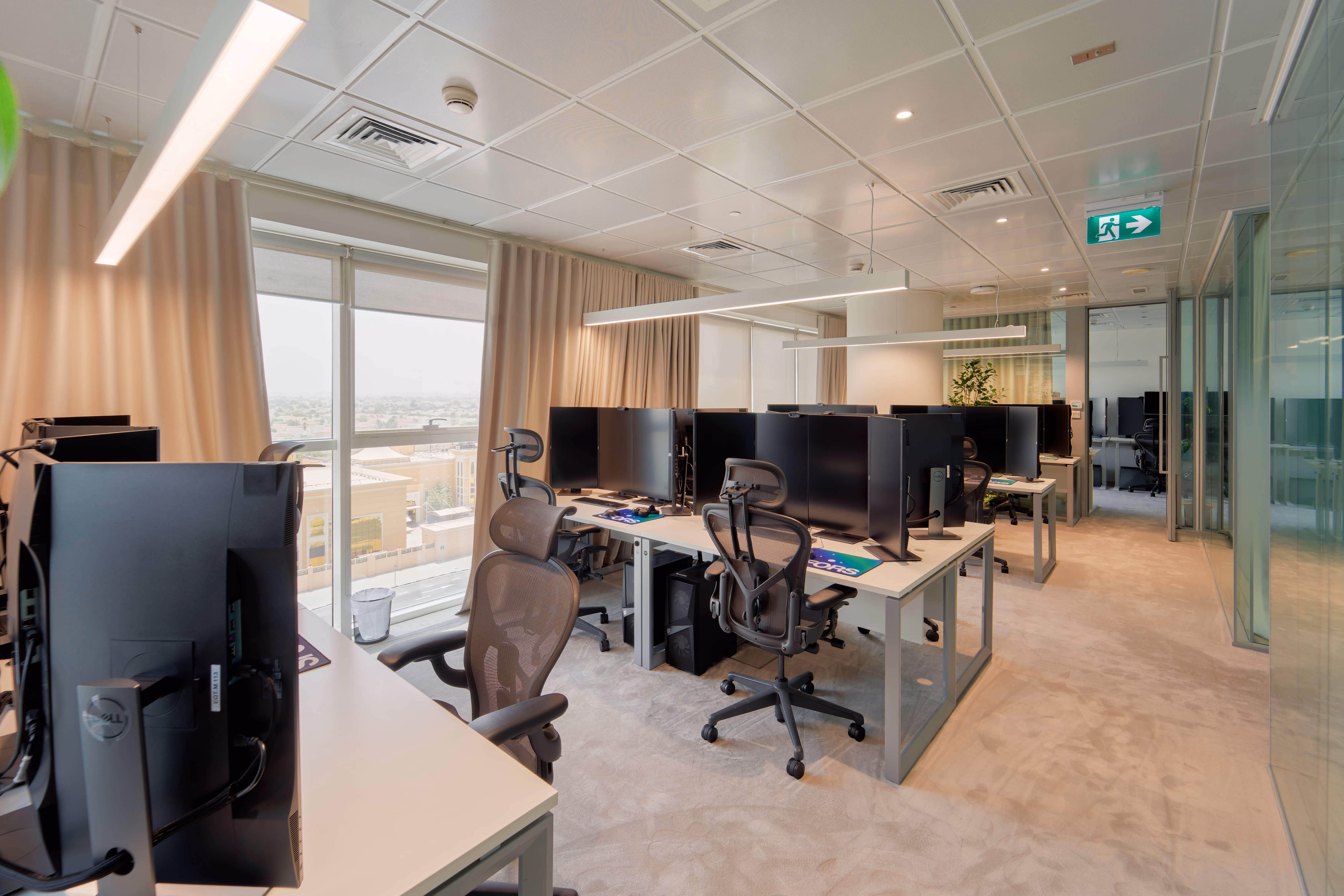 Modern office with multiple workstations, ergonomic chairs, large windows, and beige curtains.