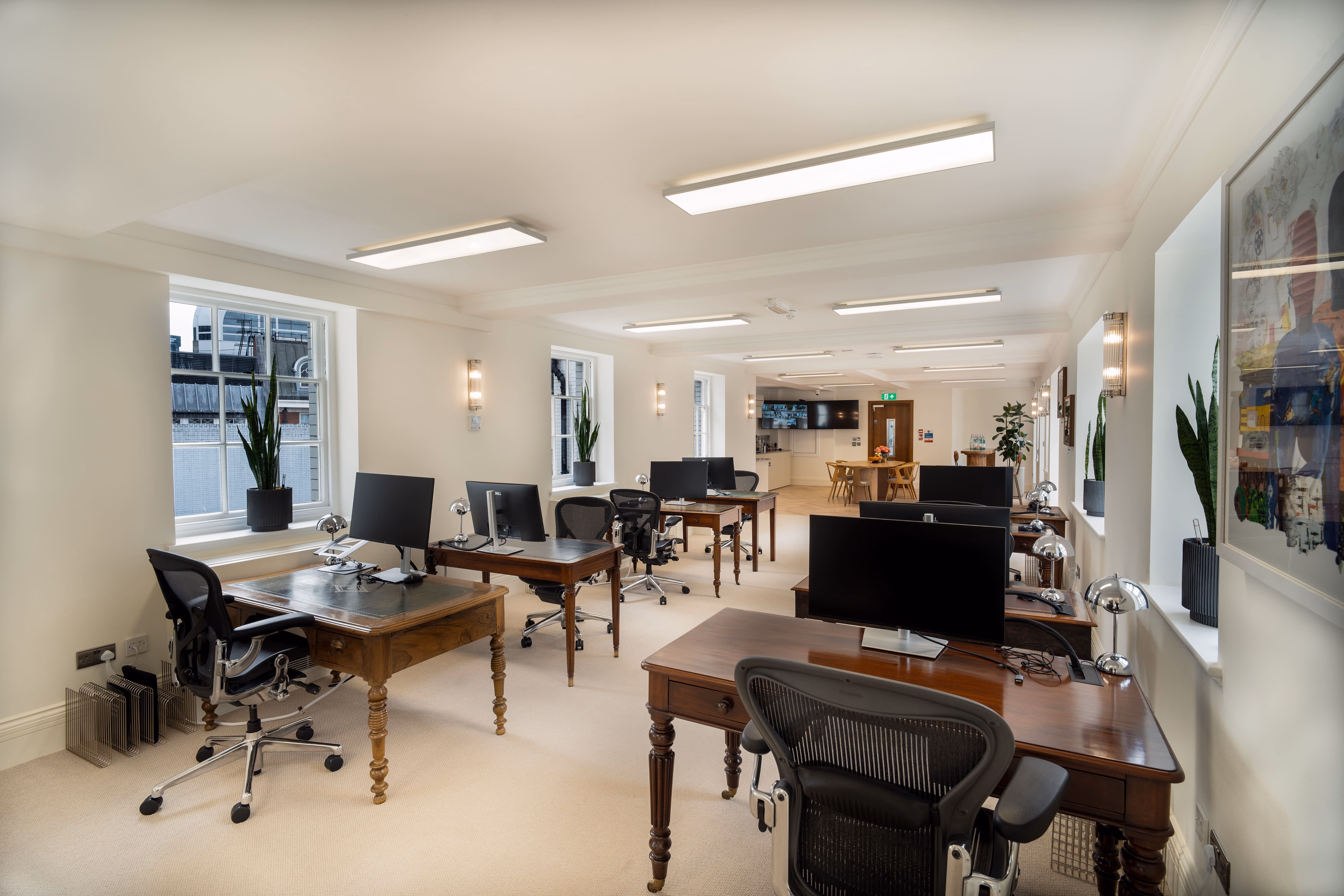 Bright office with wooden desks, black mesh chairs, computer monitors, and plants by large windows.