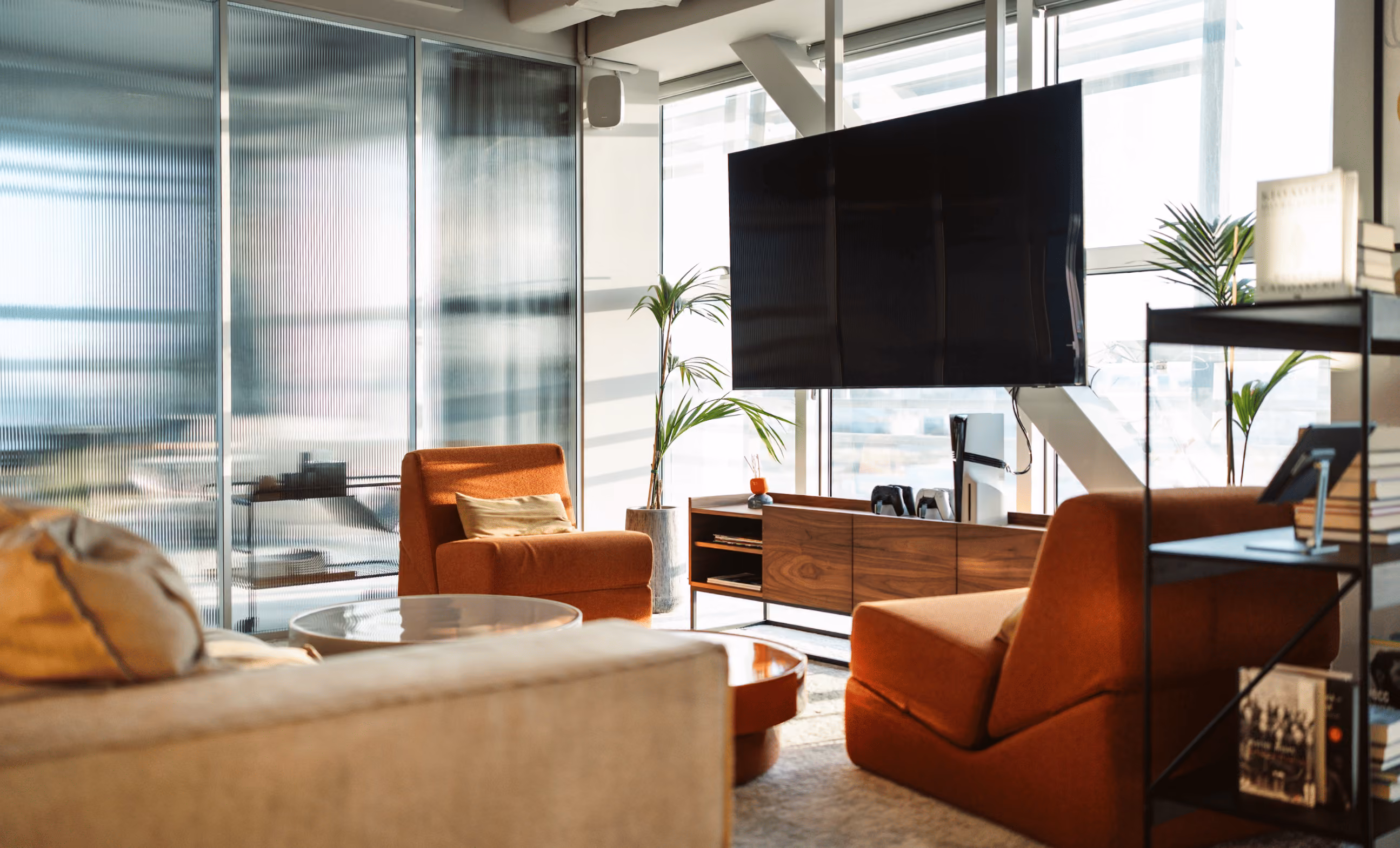 Bright office with wooden desks, black mesh chairs, computer monitors, and plants by large windows.