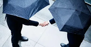 Two businessmen shake hands while holding umbrellas during a rain storm.