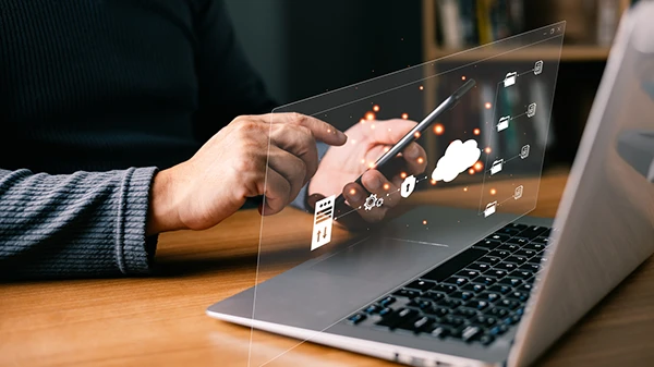 A person uses a phone above a laptop keyboard, with icons floating above the screen and their hands.