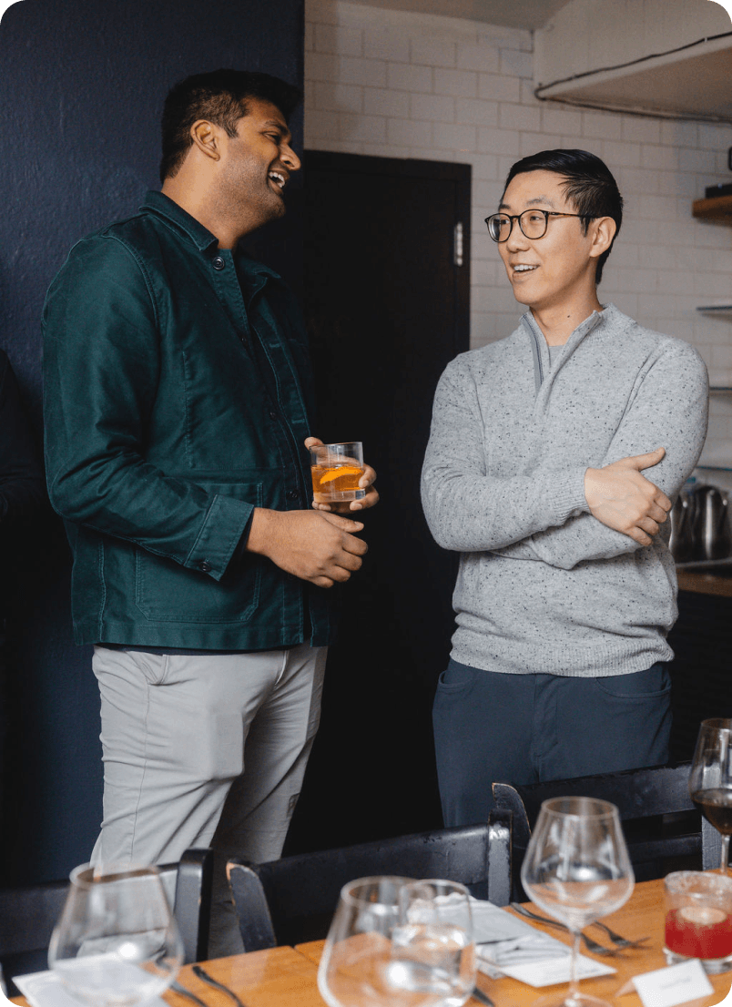 Two men standing and talking, one holding a drink, with a table set with glasses in front of them.