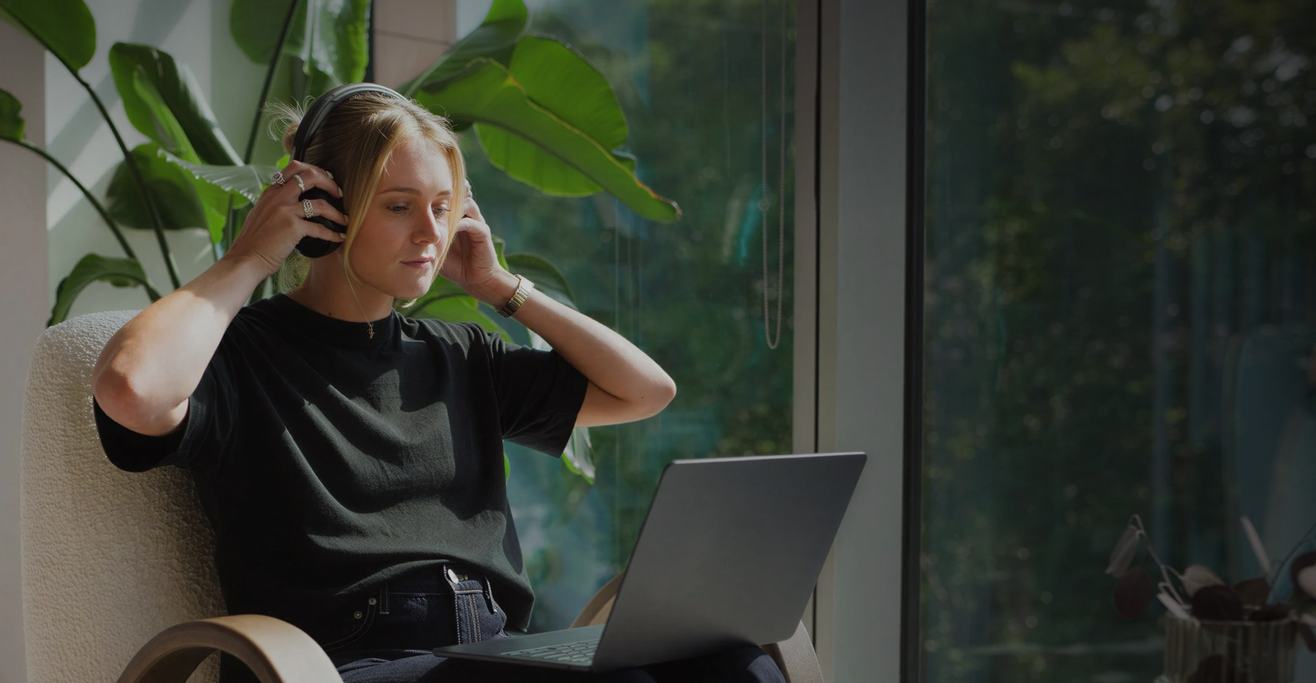 Woman wearing headphones, seated by a window with laptop on lap and large green plant behind her.