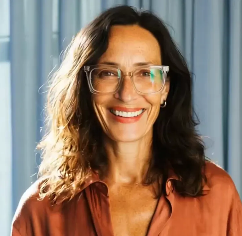 Smiling woman with wavy brown hair wearing transparent glasses and a rust-colored shirt.
