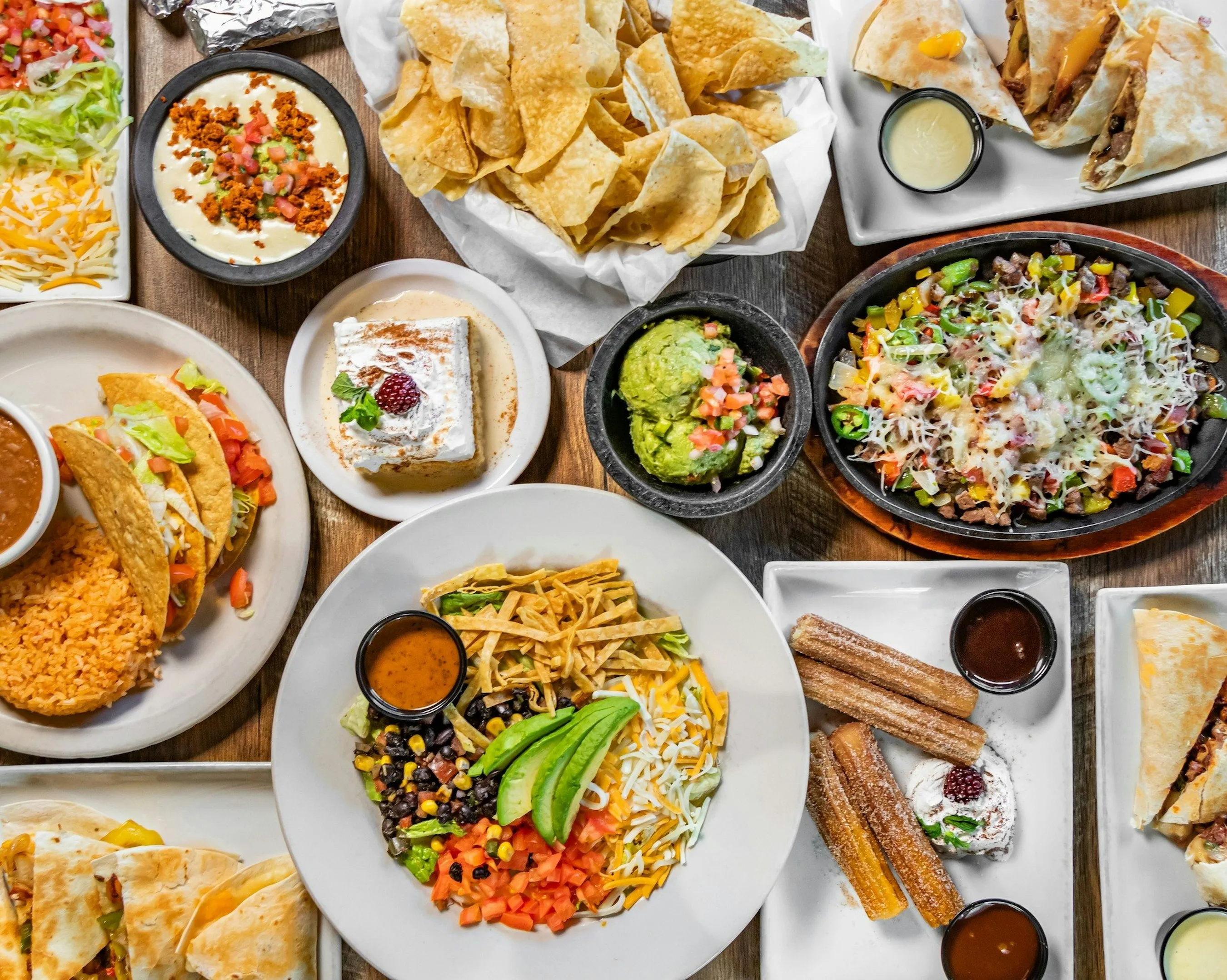 Table spread with Mexican dishes including tacos, quesadillas, chips, guacamole, salad, and churros with chocolate sauce.