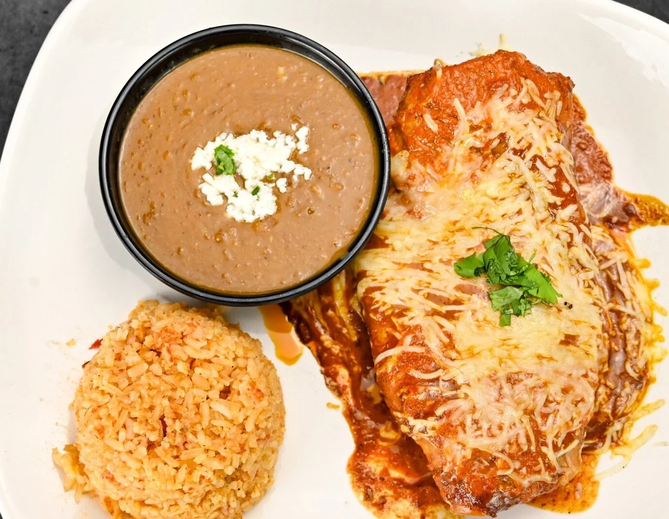 Plate with Mexican rice, refried beans topped with cheese, and a cheese-covered enchilada garnished with cilantro.