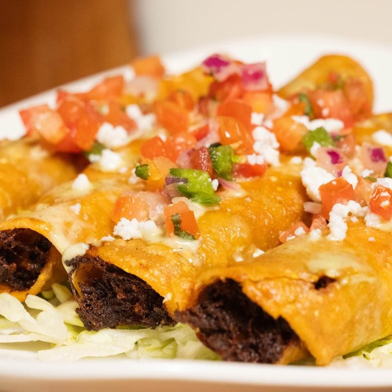 Plate of fried flautas topped with diced tomatoes, onions, peppers, and crumbled cheese on shredded lettuce.
