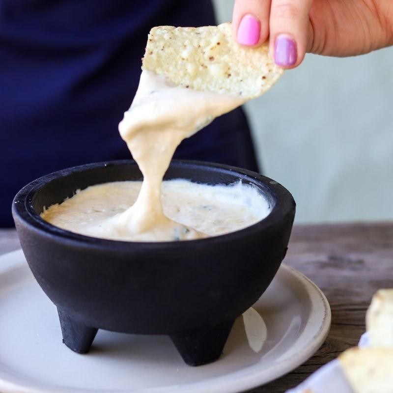 Hand with purple and pink nail polish dipping a chip into creamy white queso in a black bowl.
