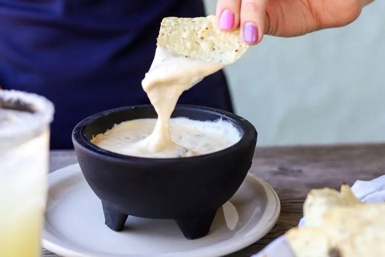 Hand dipping a tortilla chip into a black bowl of creamy white queso dip on a beige plate.