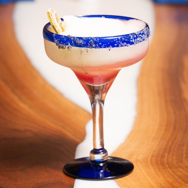 Cocktail in a glass with a blue rim and base, garnished with a citrus slice on a wooden surface.