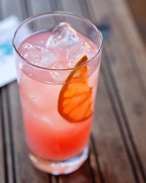 Tall glass of pink cocktail with ice and a dried orange slice on a wooden surface.