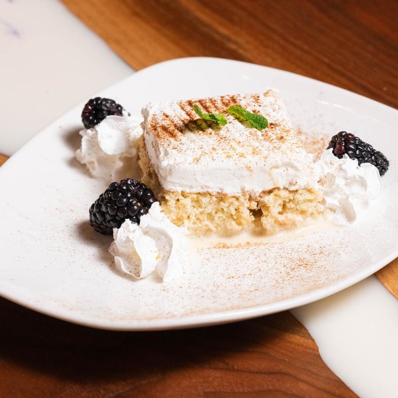 A slice of tres leches cake topped with whipped cream, mint, and blackberries on a white plate.