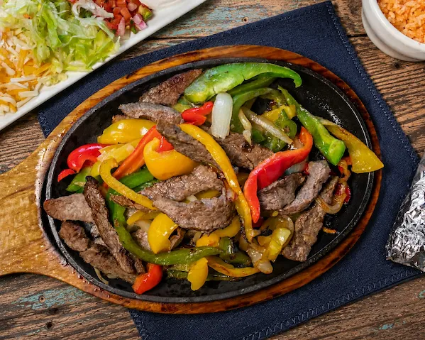 Sizzling steak fajita with grilled bell peppers and onions on a wooden platter with sides nearby.
