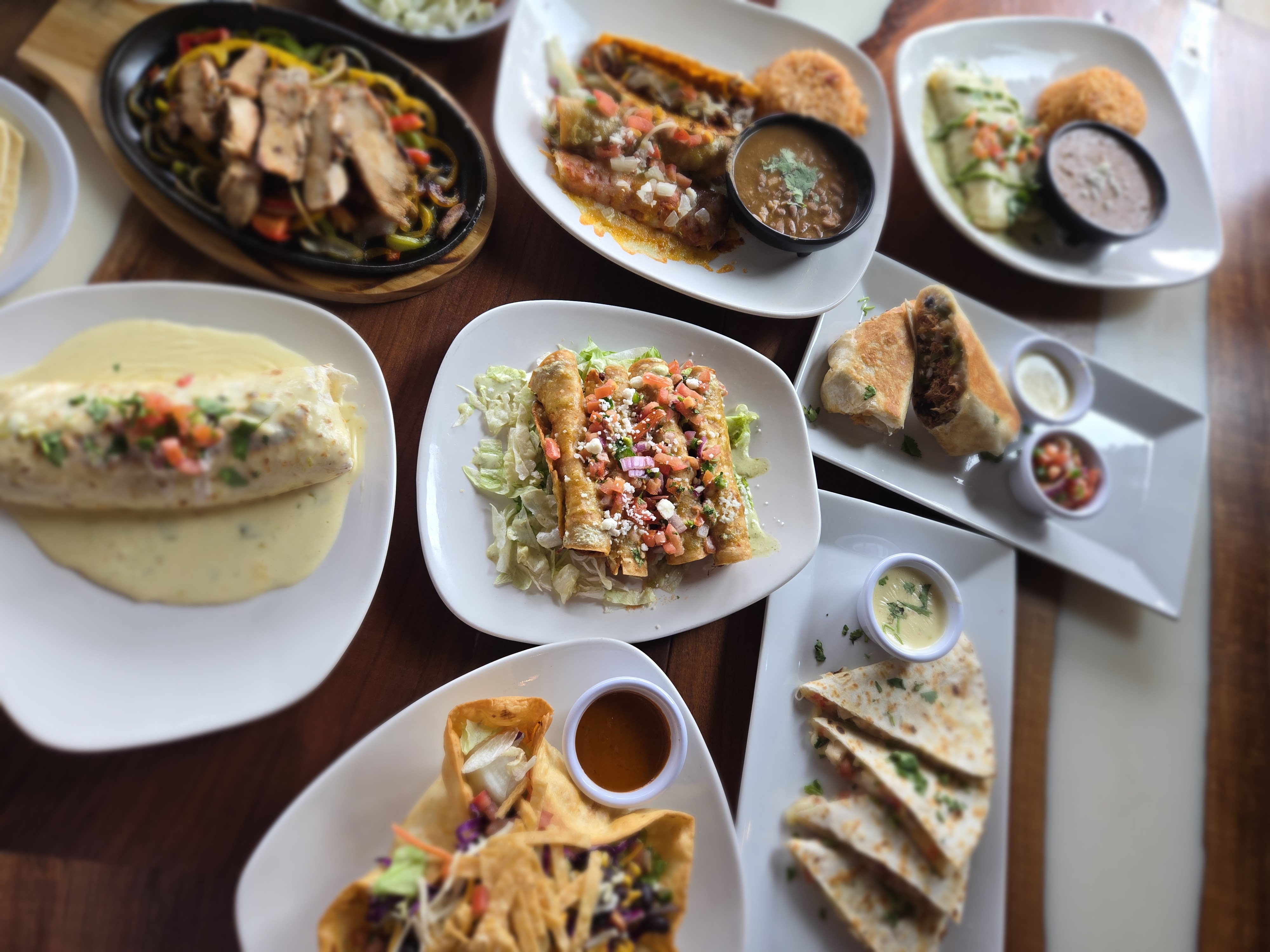 Table with various Mexican dishes including enchiladas, quesadillas, flautas, fajitas, and sides of rice and beans.