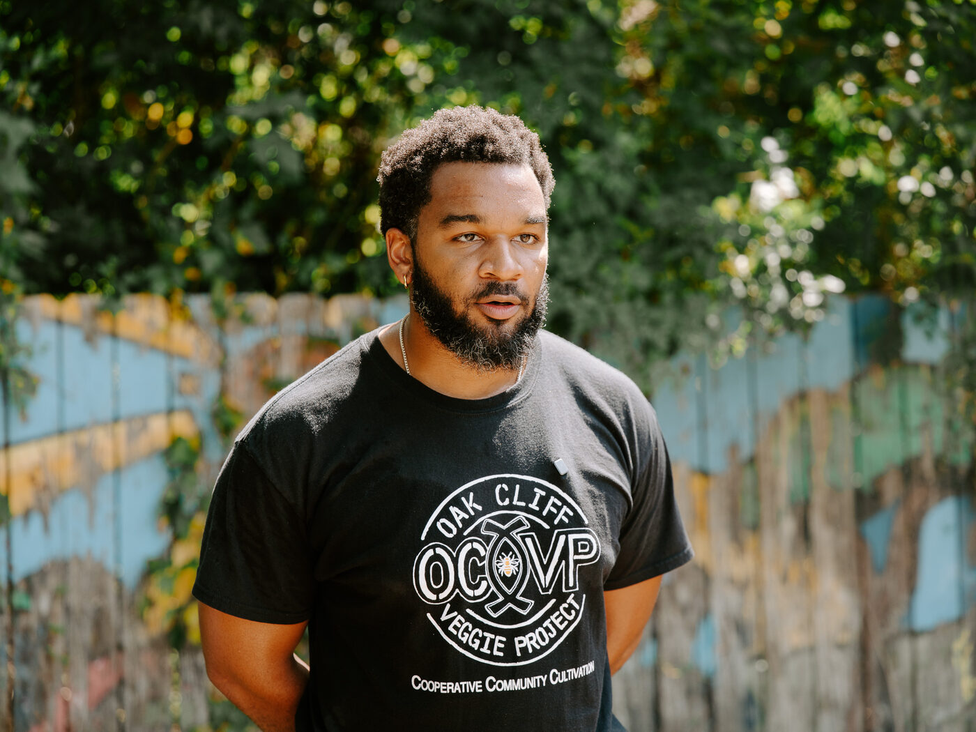 An Oak Cliff Veggie Project leader in a project t-shirt stands beside the garden's painted fence