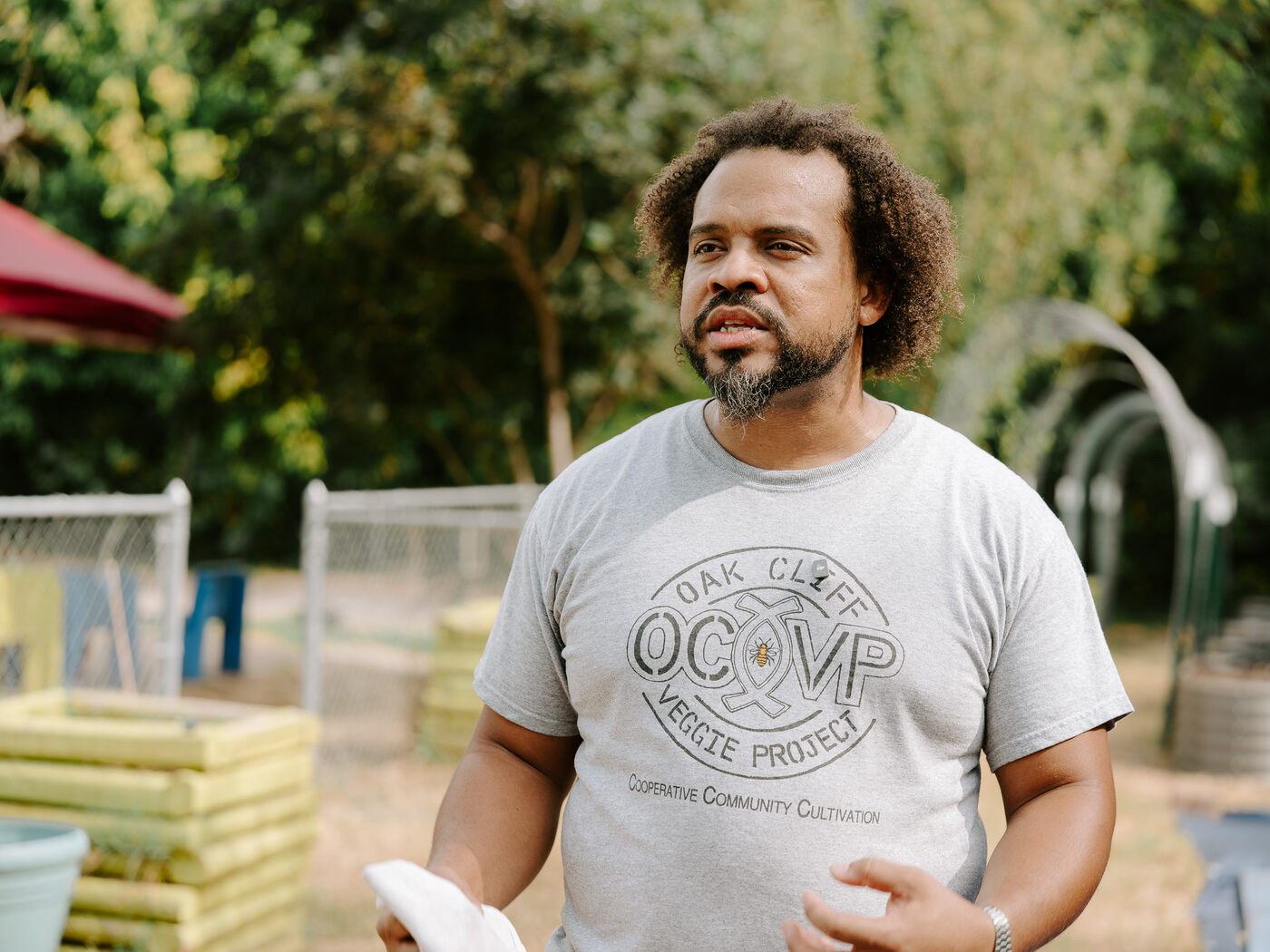 An Oak Cliff Veggie Project team member in a project t-shirt stands among the garden's arched trellises