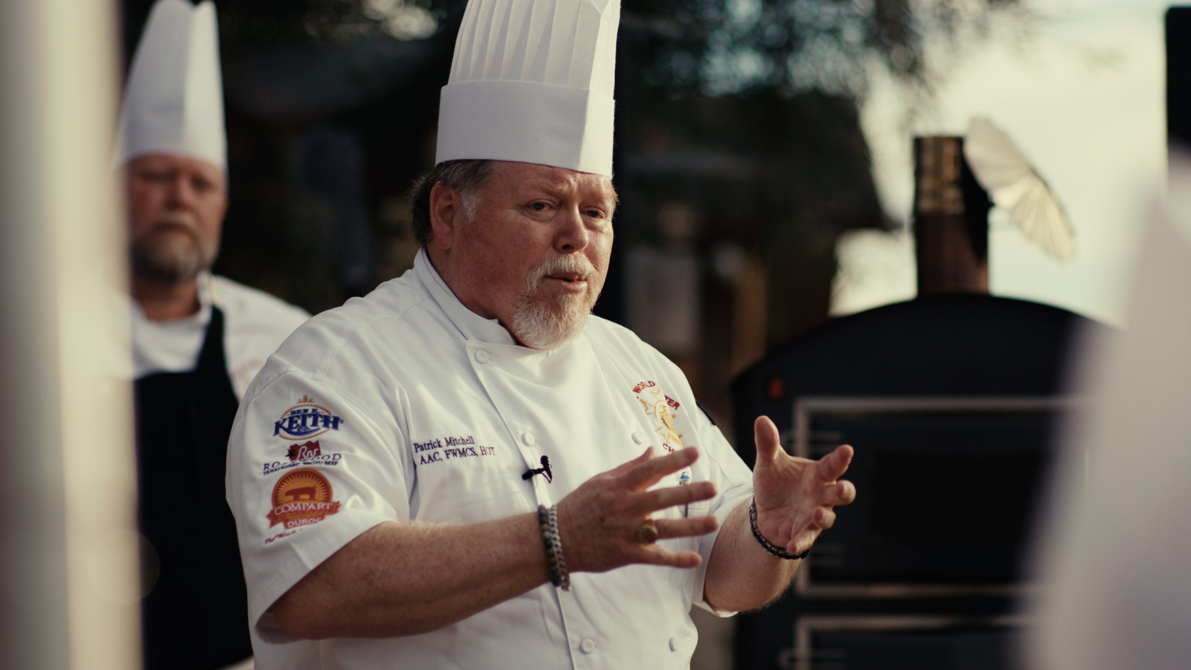 A Master Chef in whites and toque teaching at the pass in a working kitchen