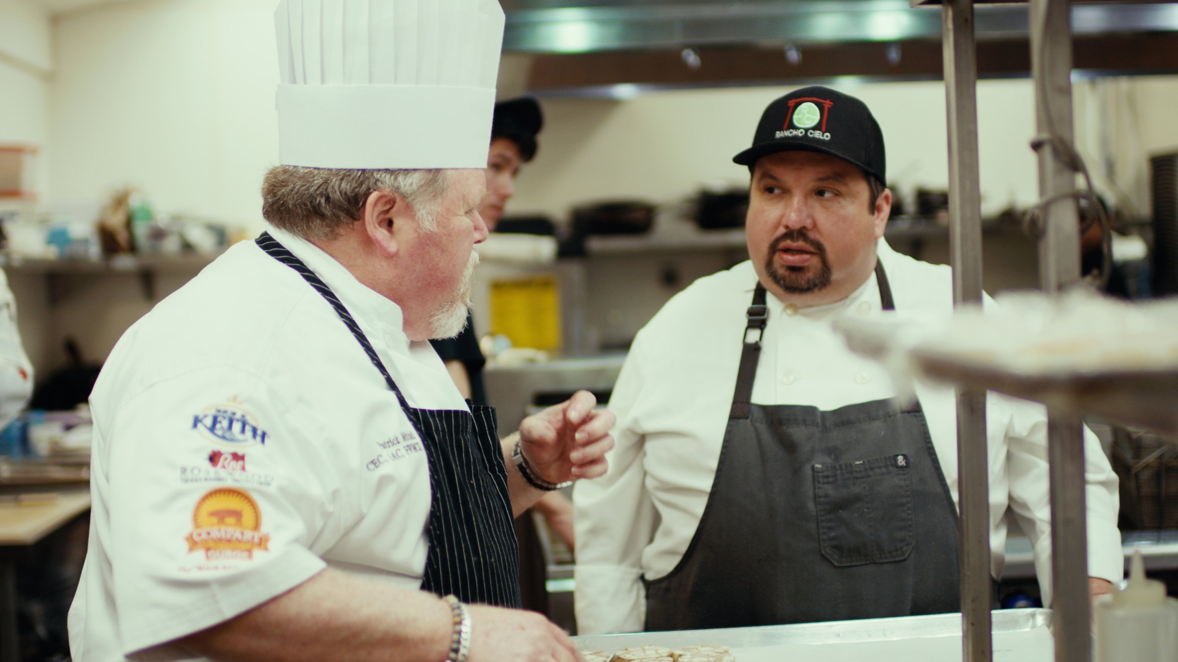 Two Master Chefs in whites talking at the pass in a working kitchen