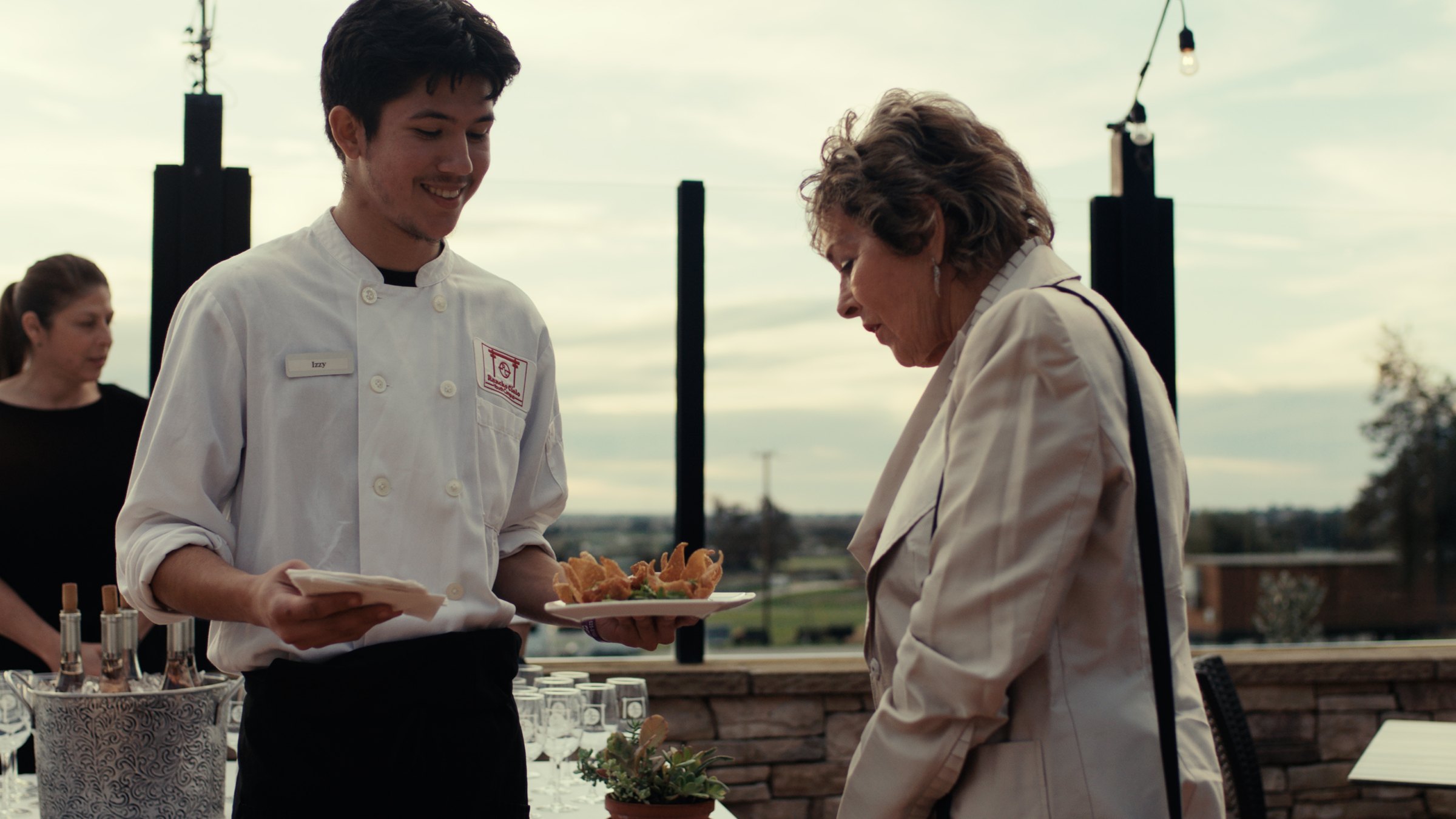 A young chef in whites serving a guest at an outdoor chapter event