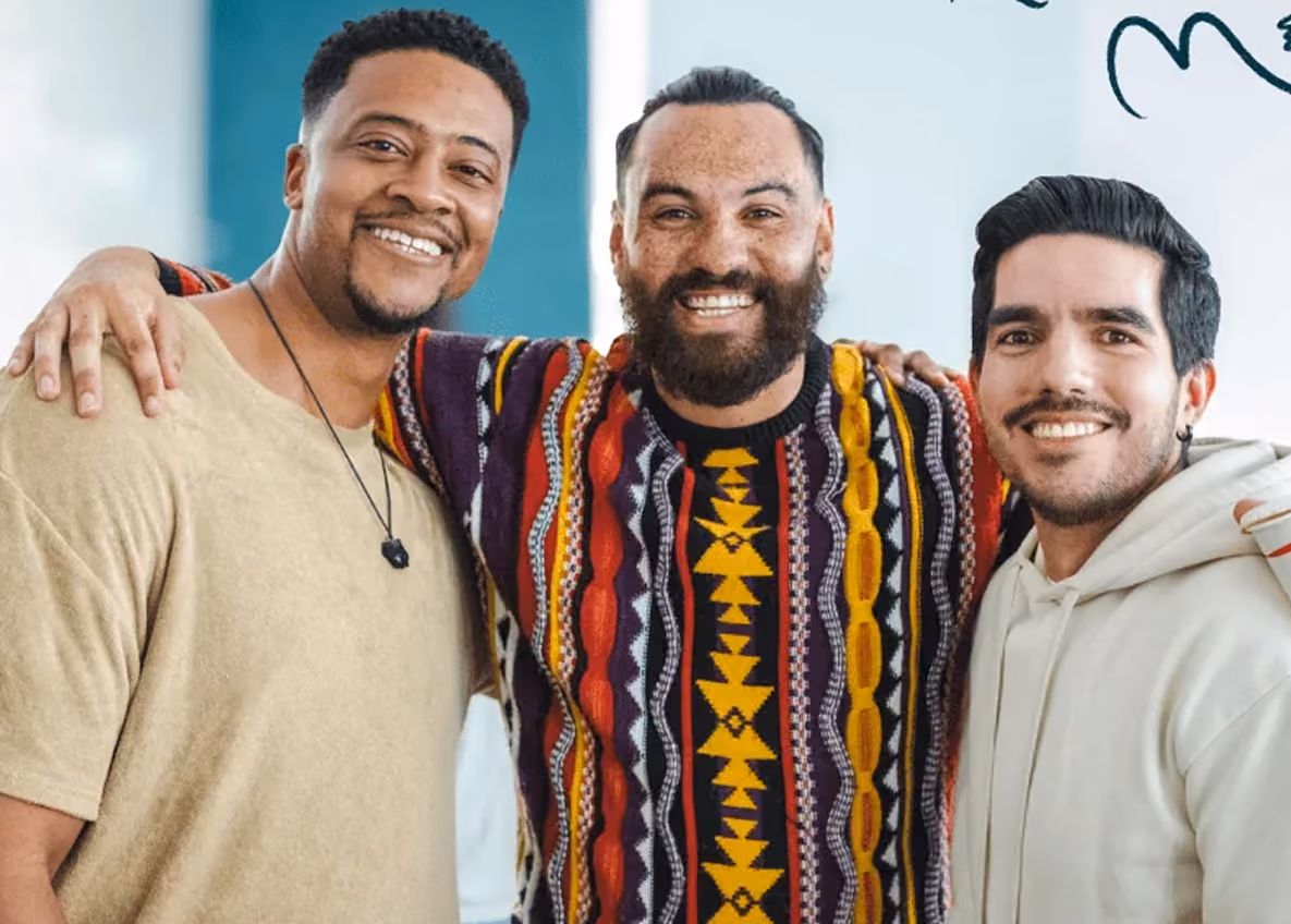 Photo of three smiling men with arms around each other, one wearing a colorful patterned poncho.