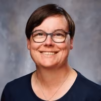 Headshot photo of a smiling woman with short brown hair and glasses against a gray background.