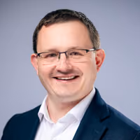 Headshot portrait of a smiling man wearing glasses and a navy suit jacket.