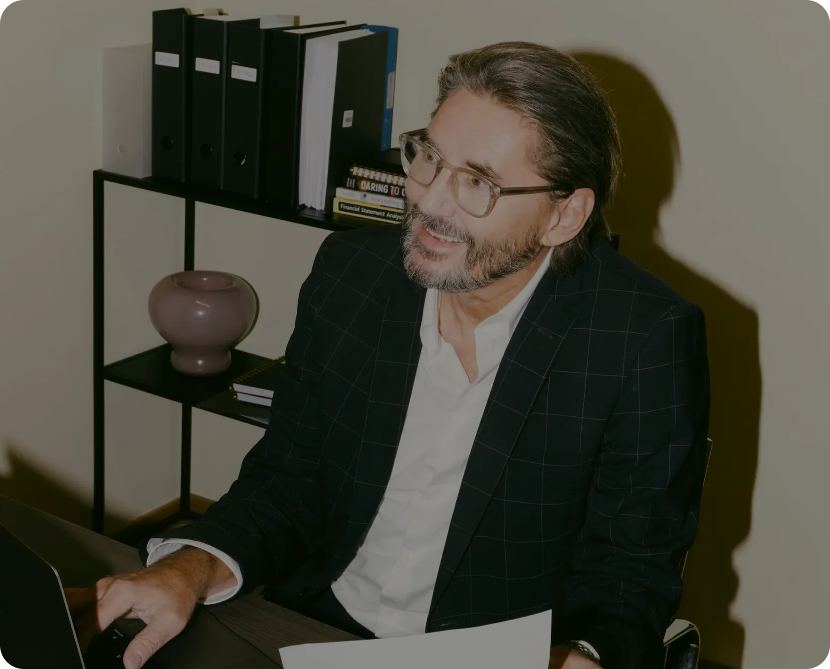 Headshot of a man wearing glasses and a blazer working at a laptop in an office with bookshelves.