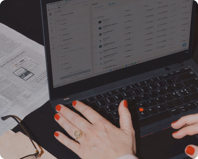 Photo of hands typing on a laptop displaying a list of contact records in the Surfe interface, with papers and glasses nearby.