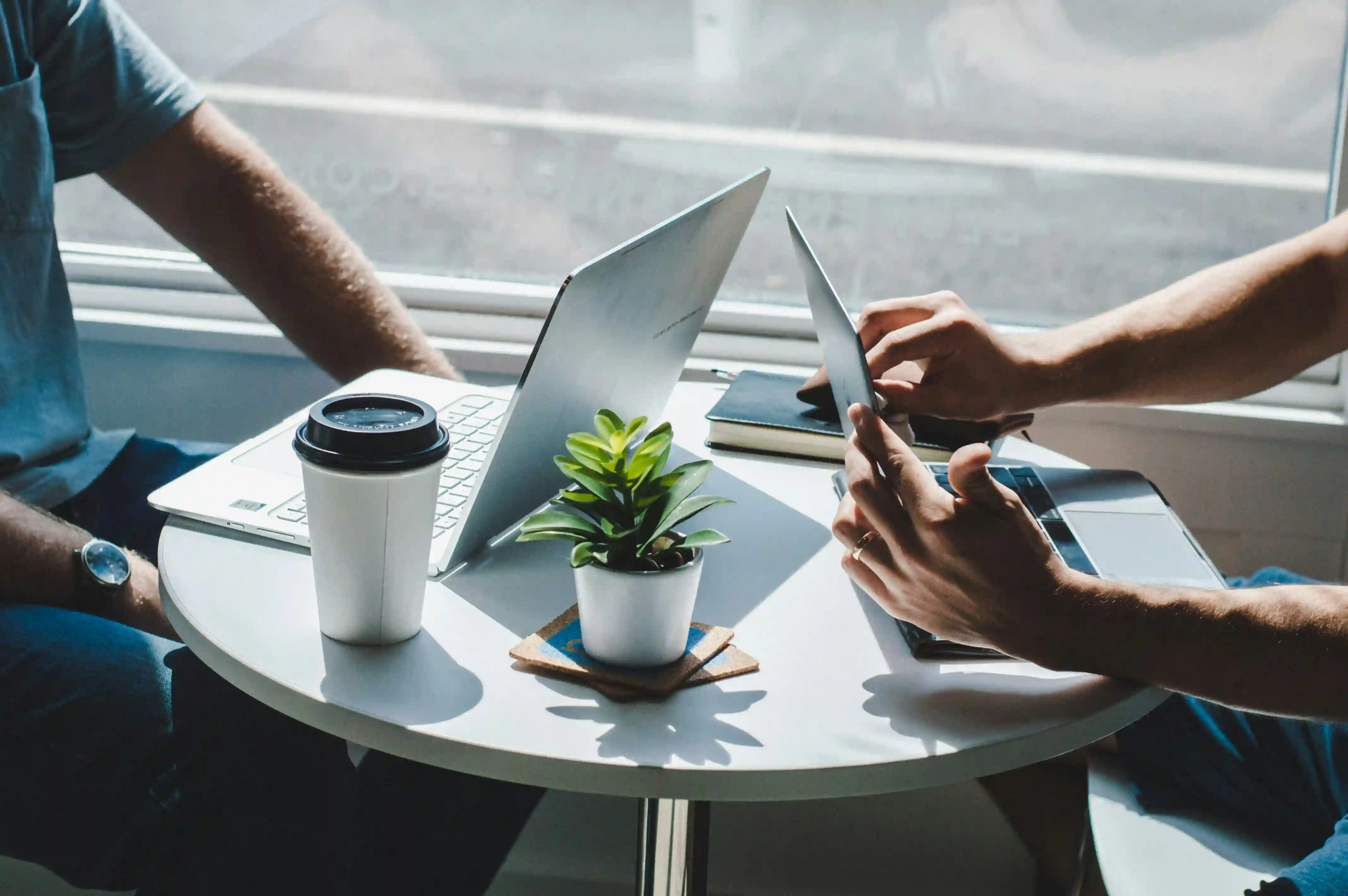 Two people sitting at a white round table with laptops, a tablet, a plant, and a coffee cup.