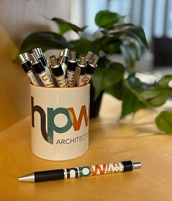 Mug with "hpw architecture" logo holding pens on a wooden table. One pen lies beside it.