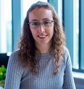 A photo of Emma Jones, Architect. Smiling woman with glasses and curly hair in a gray sweater sits indoors. Bright blue windows in the background add a calm ambiance.