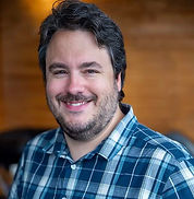Photo of Huw Jones, Architectural Designer. Smiling man in blue plaid shirt against a blurred wooden background. Warm lighting creates a friendly, relaxed atmosphere.