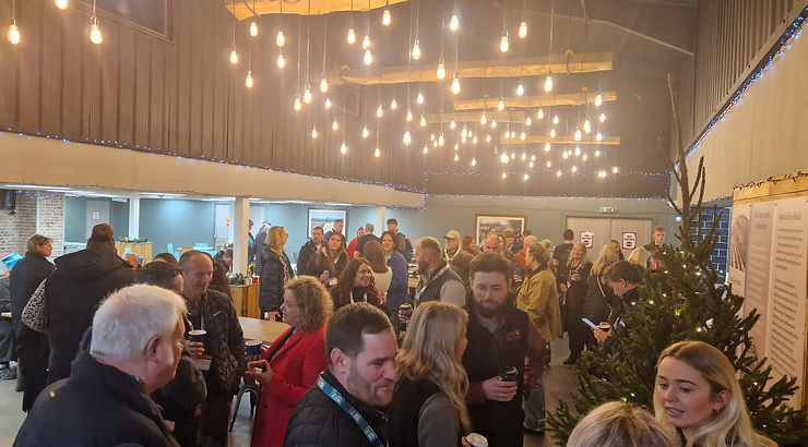 People mingling at a festive indoor gathering with string lights overhead and a Christmas tree. Mood is lively and social.