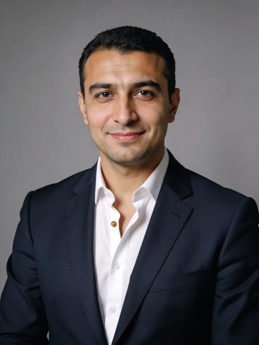 Smiling man with short dark hair wearing a white shirt and dark suit jacket against gray background.