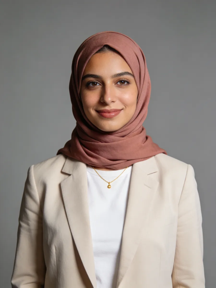 Smiling woman wearing a brown hijab, beige blazer, white shirt, and gold necklace against gray background.