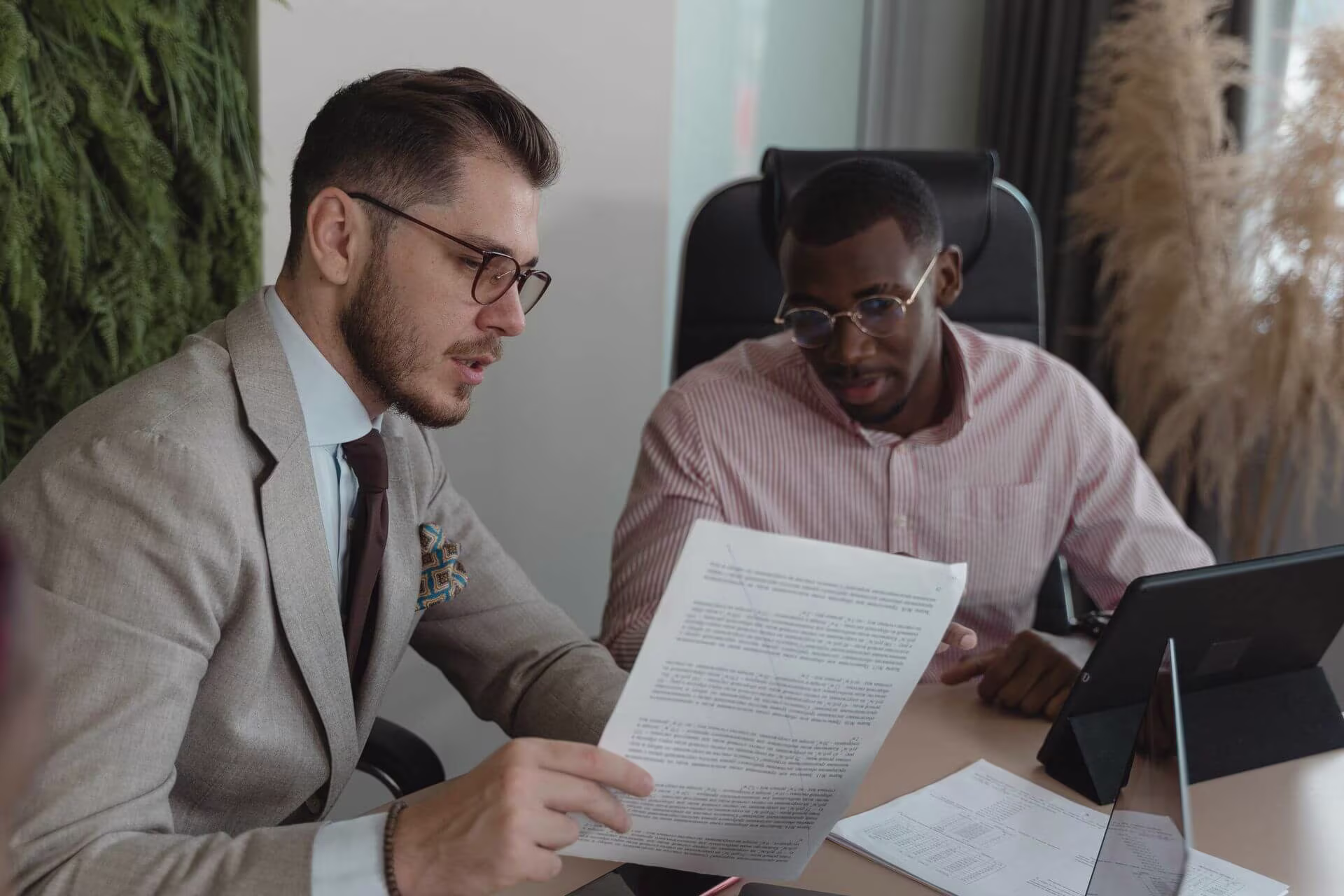Two men in glasses reviewing a document together at a desk with a tablet and papers.