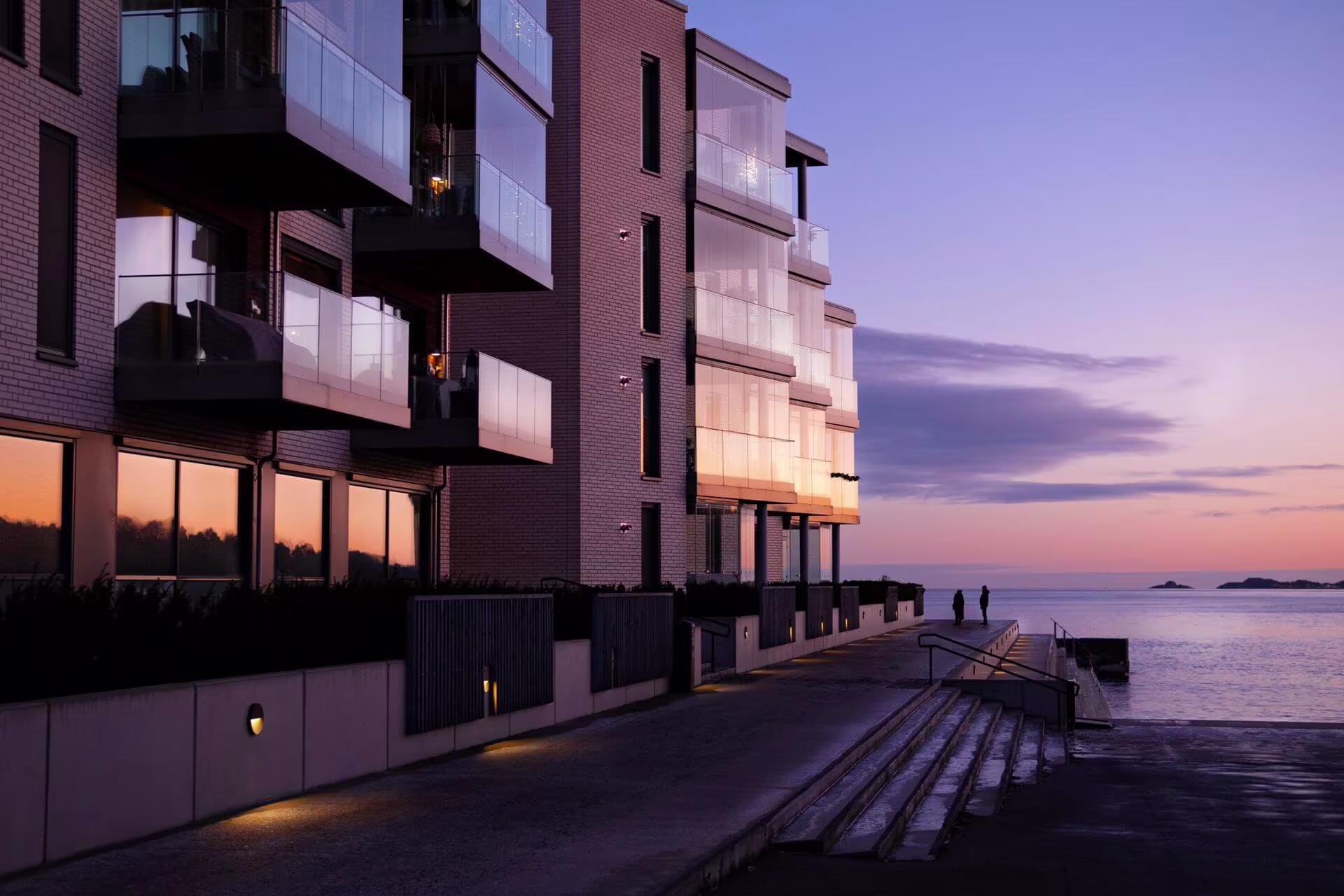 Modern apartment building with glass balconies by the water at sunset with two people on the pier.