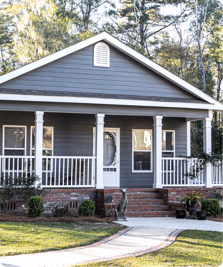 Front view of a gray house with white porch railing, brick steps, green bushes, and a dog statue.