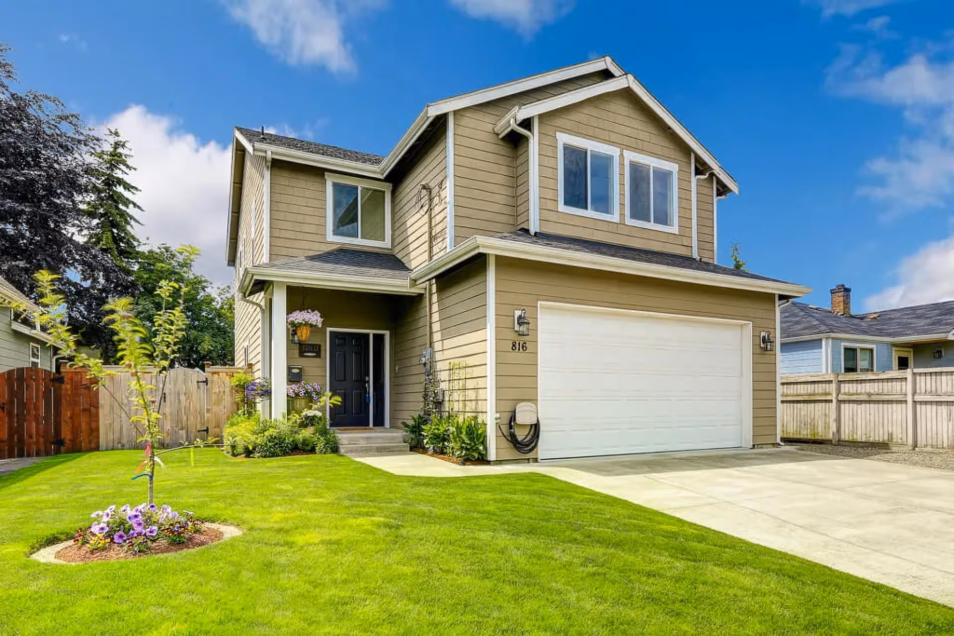 Two-story beige house with a black front door, white garage door, green lawn, and flower bed.