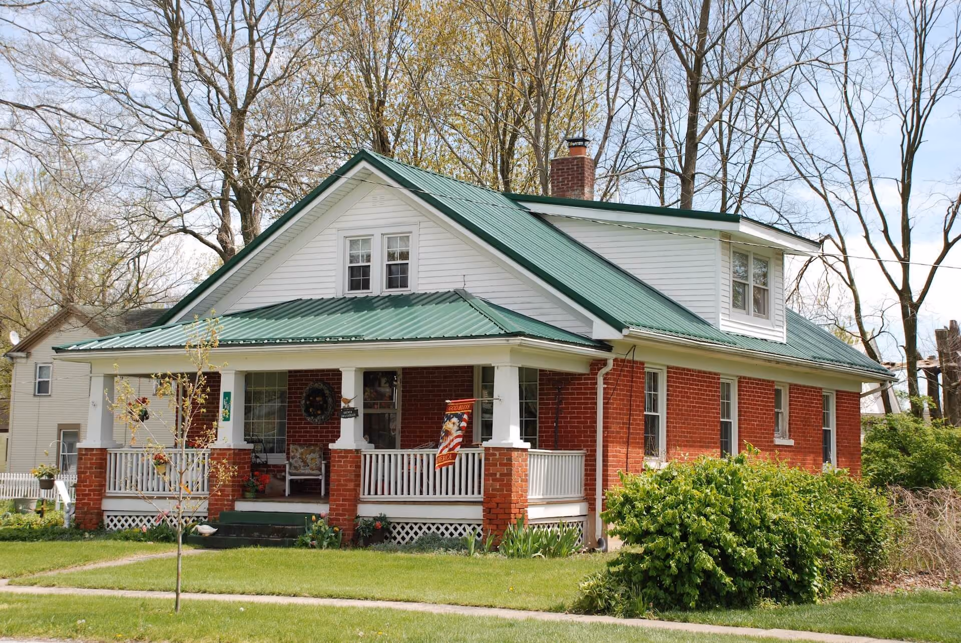 Red brick house with white trim, green metal roof, front porch, and trees in the background.