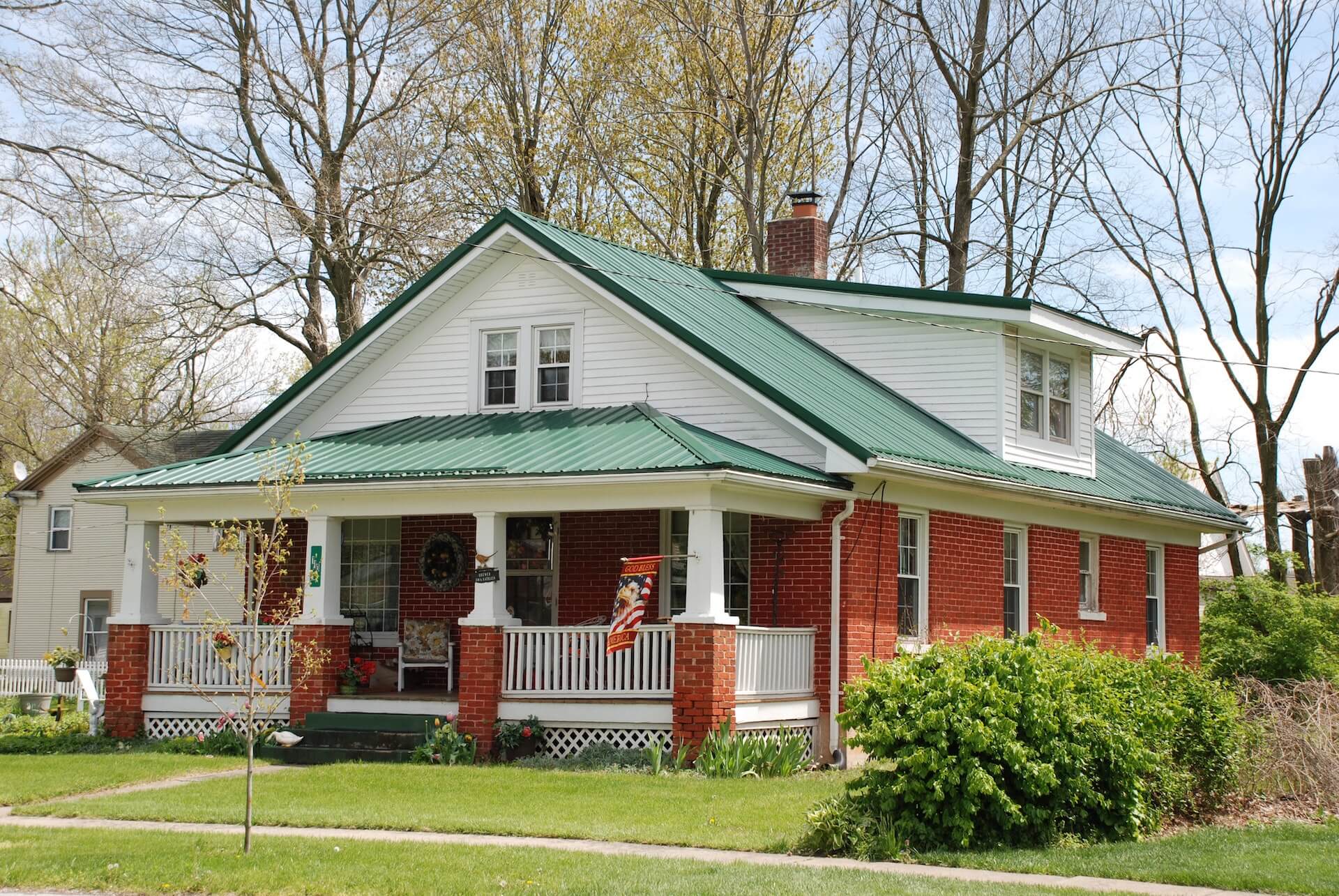 Red brick house with white trim, green metal roof, front porch, and trees in the background.
