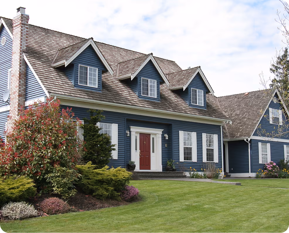 Blue house with wooden roof, red door, white window shutters, and a well-kept lawn with shrubs.