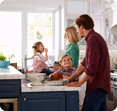 Family of four in a kitchen with father holding a tray of cooked chicken and mother with children near the sink.