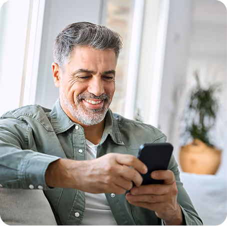 Smiling middle-aged man using a smartphone while sitting on a couch near a window at home.