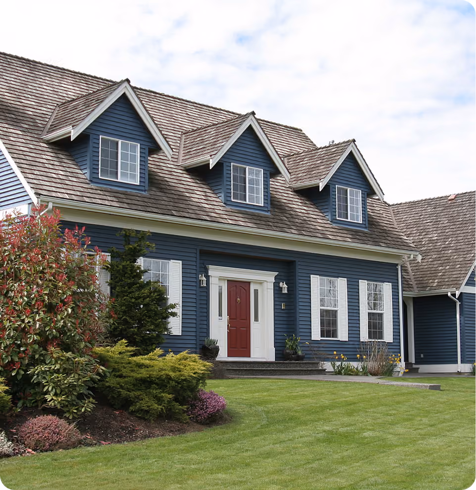 Two-story blue house with white shutters, a red front door, and a green lawn with bushes.